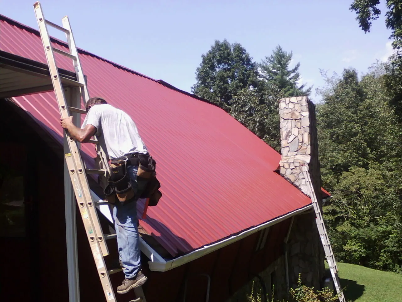 Roofer on a ladder beside a red metal roof and stone chimney — Soffit & Fascia Repair by Roofing Brothers in Knights Landing